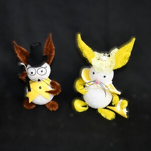 May include: Two handmade bunny figurines, one with brown fur and a top hat, the other with yellow fur and a yellow hat. Both bunnies have white faces and black eyes.
