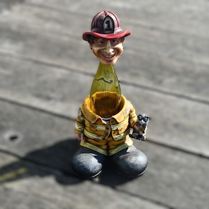 Pode incluir: Uma estatueta decorativa de bombeiro com um capacete vermelho com o número um, um casaco amarelo e botas pretas. A cabeça está presa ao gargalo de uma garrafa de vidro transparente e a figura segura um pequeno animal preto e branco.