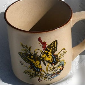 May include: Beige ceramic mug with a brown rim and handle. The mug features a colorful butterfly design with yellow, black, and blue accents, along with floral and house illustrations. The mug is suitable for hot or cold beverages.
