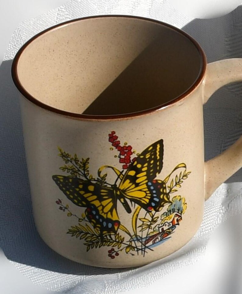 May include: Beige ceramic mug with a brown rim and handle. The mug features a colorful butterfly design with yellow, black, and blue accents, along with floral and house illustrations. The mug is suitable for hot or cold beverages.