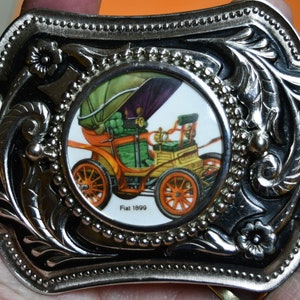 Puede incluir: Hebilla de cinturón de metal plateado con un diseño floral ornamentado. El centro presenta una imagen circular de un coche Fiat 1899 vintage naranja y verde. La hebilla tiene forma rectangular con un borde decorativo.