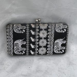 May include: A rectangular black and white wallet featuring a repeating pattern of elephants and floral motifs. The wallet has a silver-toned metal kiss-lock clasp at the top center, providing a secure closure for the structured, fabric-covered accessory.