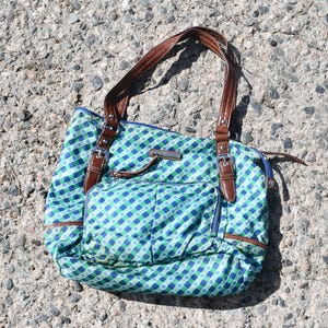 Könnte beinhalten: Eine blau, grün und weiß karierte Tragetasche mit braunen Ledergriffen und -besatz. Die Tasche hat eine Vordertasche mit Reißverschluss und einen blauen Reißverschluss. Die Tasche liegt auf einer grauen, strukturierten Oberfläche.