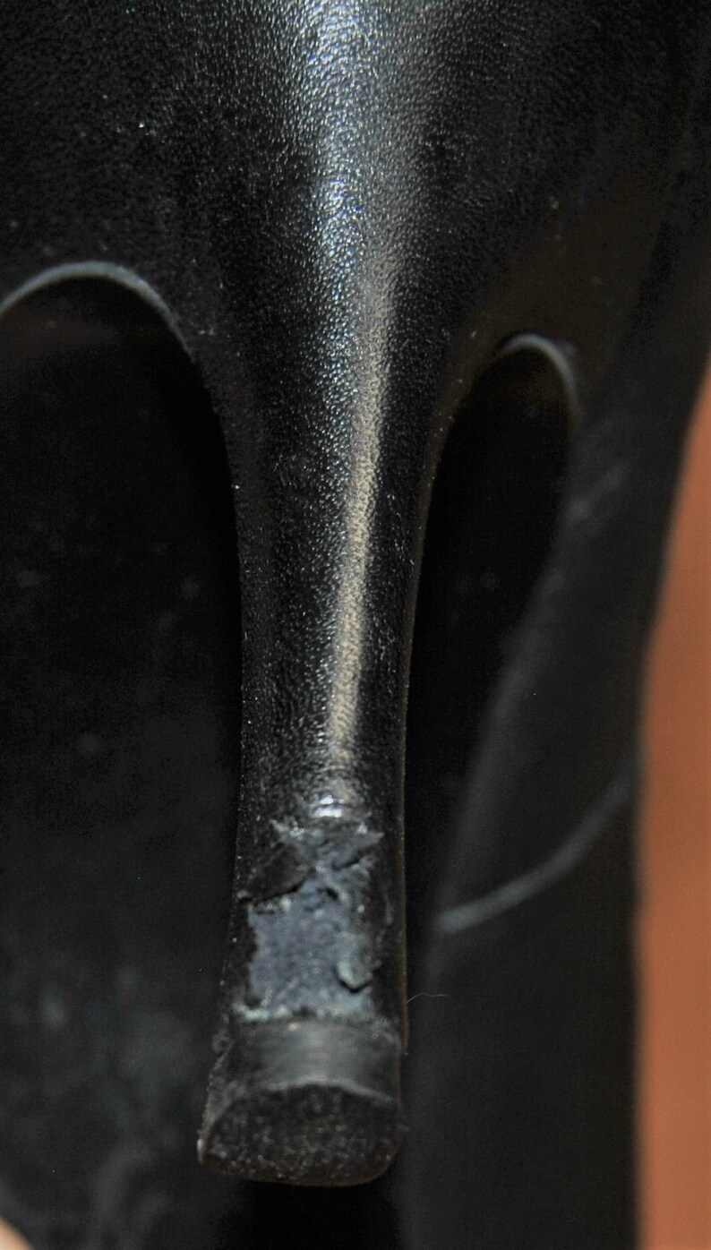 May include: Close-up of the bottom of a black leather heeled shoe. The heel is worn and has a small tear in the leather.