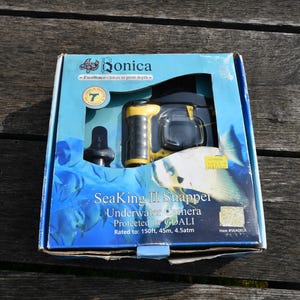 May include: A Bonica SeaKing II Snapper underwater camera in a blue cardboard box. The packaging features a yellow and black camera, fish imagery, and text stating it is rated to 150ft, 45m, and 4.5atm. The box has a clear plastic window.