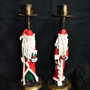 May include: Two Santa Claus candle holders made of clay. Each Santa is wearing a red suit with white trim, a white beard, and a red hat. They are holding a staff and a gift. The candle holders are made of metal and have a gold finish.