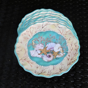 May include: A stack of decorative plates with a coastal theme. Each plate features a scalloped edge in turquoise, surrounding a central design of seashells, starfish, and seahorses in shades of beige, turquoise, and coral.