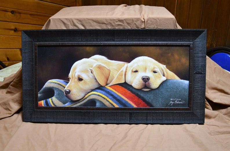 May include: Framed painting of two yellow Labrador Retriever puppies sleeping on a colorful blanket. The painting is signed by the artist, "Jay Johnson".
