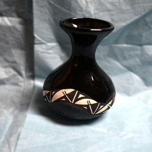 Puede incluir: Un jarrón de cerámica negro con un diseño geométrico blanco. El jarrón tiene un cuerpo redondo y un cuello estrecho.