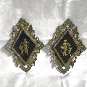 May include: Two antique wall plaques with ornate, gold-toned frames. Each plaque features a black diamond-shaped center with a gold relief of a woman and child. The frames have a detailed, scrollwork design.