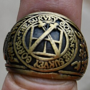 May include: A gold-colored ring with the inscription "Corps of the Survey" and a graphic of two crossed tools. The ring is worn on a finger.
