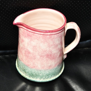 May include: A pink and green ceramic pitcher with a speckled glaze. The pitcher has a wide mouth and a handle.
