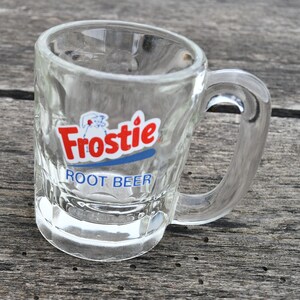 May include: A clear glass mug featuring a textured exterior and a sturdy handle. The front of the mug displays the red and white Frostie logo above the blue text ROOT BEER. The glass has a thick base and a smooth rim, designed for serving beverages.