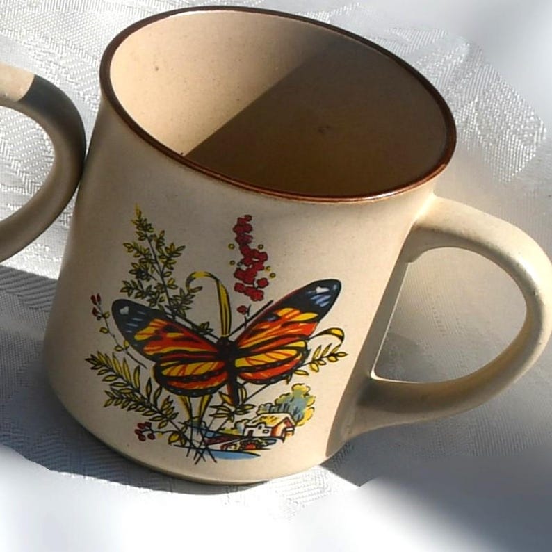 May include: Beige ceramic mug with a brown rim and handle. The mug features a colorful butterfly design with red, orange, yellow, and black wings, surrounded by green foliage and a small house illustration.