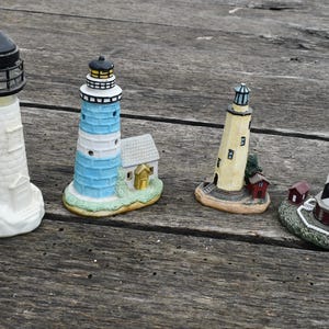 May include: Four decorative lighthouse figurines in various designs. One is white, another blue and white, a third yellow, and the last is black and white striped. Each has a detailed top and sits on a base.