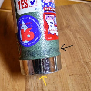 May include: A green and silver insulated tumbler with various stickers, including "YES" and "BUY AMERICAN." The tumbler features a chrome-colored base and is resting on a wooden surface. The stickers include "Local Union 16 Omaha, Nebraska" and "IBEW Local 1077."