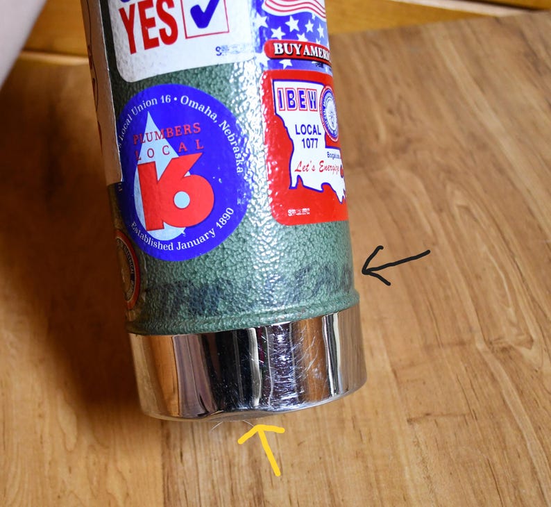 May include: A green and silver insulated tumbler with various stickers, including "YES" and "BUY AMERICAN." The tumbler features a chrome-colored base and is resting on a wooden surface. The stickers include "Local Union 16 Omaha, Nebraska" and "IBEW Local 1077."