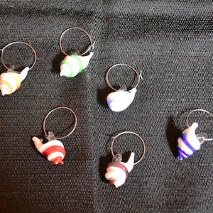 Könnte beinhalten: Sechs bunte gläserne Schnecken-Wein-Anhänger mit silbernen Ringen. Die Schnecken sind weiß mit verschiedenen farbigen Streifen. Die Farben umfassen Grün, Orange, Rot, Blau und Lila.