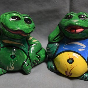 Könnte beinhalten: Zwei handbemalte Keramik-Froschfiguren. Ein Frosch ist grün mit weißen und grünen Details, der andere ist grün mit blauen, gelben und weißen Details.