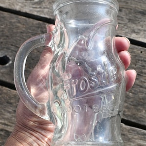 May include: Clear glass mug with a handle, embossed with the word "Frostie" and "Root Beer". The mug has a rounded body and a flared rim. The mug is held by a person.