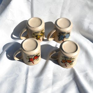 May include: Four cream-colored ceramic mugs with curved handles, each adorned with a different butterfly design in shades of red, yellow, and blue. The mugs are arranged on a white, textured surface.