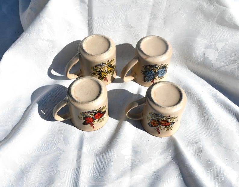May include: Four cream-colored ceramic mugs with curved handles, each adorned with a different butterfly design in shades of red, yellow, and blue. The mugs are arranged on a white, textured surface.