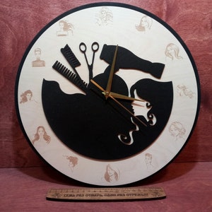 May include: A round wooden clock with a black face featuring a silhouette of a woman's hair and styling tools, including a hairdryer, scissors, and combs. The clock face is surrounded by a white ring with 12 images of women's hairstyles.