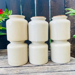 Peut inclure: Six pots en céramique blanc cassé avec des bouchons en liège sont empilés sur une surface en bois. Les pots ont une forme cylindrique avec une base plus large et un sommet plus étroit. Le mot "LABBY" est imprimé au bas des pots. Une fleur rouge et des feuilles vertes sont en arrière-plan.