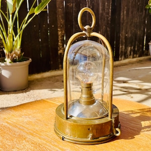 May include: A vintage brass lamp with a glass dome and a light bulb inside. The lamp is sitting on a wooden table.