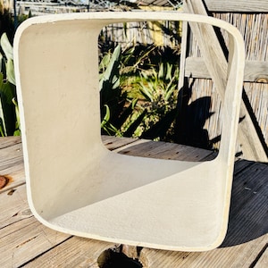 Peut inclure: Un cube décoratif carré, ouvert sur les côtés, de couleur blanc cassé. Le cube est fait d'un matériau solide avec des coins arrondis. Le cube est posé sur une surface en bois, avec de la verdure et une clôture en bois en arrière-plan.