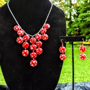 May include: A red beaded necklace and matching earrings set. The necklace and earrings feature large, round red beads with white polka dots. The jewellery is displayed on a black mannequin and stand against a green background.
