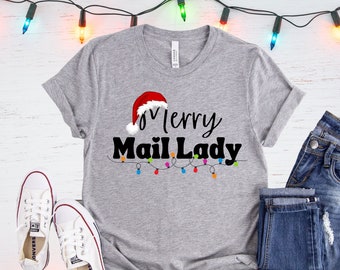 May include: A heather gray t-shirt with the text "Merry Mail Lady" in black script, a Santa hat, and a string of colorful lights. White canvas sneakers and blue jeans are also in the image. Festive holiday apparel.