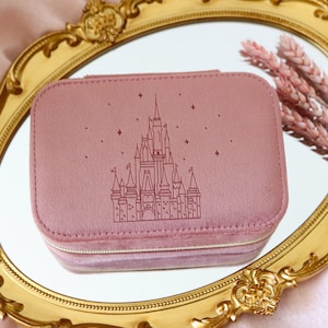 May include: A pink jewelry travel case with a castle design and stars. The case is rectangular with a zipper closure and is resting on a gold-framed mirror. Decorative pink flowers are next to the case.