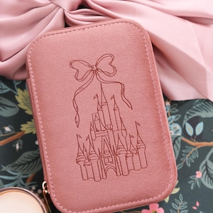 May include: A dusty rose-colored, rectangular jewelry case with a castle design and bow. The case has a gold zipper and is set against a floral background with a large pink bow. A lit candle is in the lower left corner.
