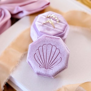 May include: A lavender velvet ring box with a hexagon shape and a seashell design. The box is open and has a ring inside.