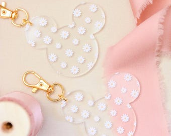 May include: Two clear acrylic keychains in the shape of Mickey Mouse's head, adorned with white daisy patterns. Each keychain has a gold-colored clasp. The background features pink ribbons and a light-colored surface.