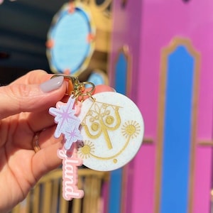 May include: A keychain featuring a white circular charm with gold line art of a stylized face, a light purple glittery star-shaped charm, and a pink rectangular charm with the white text land, all attached to a gold-toned metal ring.
