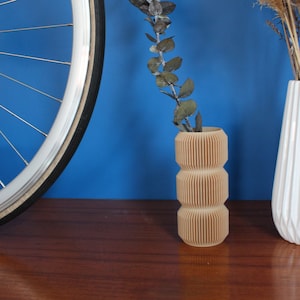 Op de afbeelding: Een houten vaas met een geribbeld ontwerp, met gedroogde eucalyptus takken. De vaas staat op een houten tafel naast een witte keramische vaas met gedroogde grassen.