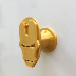 Puede incluir: Un gancho de pared de color dorado impreso en 3D con forma de la cara de Iron Man. El gancho está unido a una base redonda.