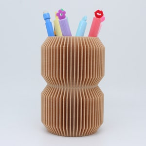 May include: A brown cardboard pencil holder with a ribbed design. The holder is filled with colourful pens and pencils, including a pink pen with a red lip topper, a light blue pen, a yellow pen, and a purple pen with a flower topper.