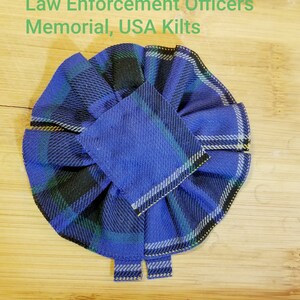 Pode incluir: Uma roseta de tecido xadrez azul, preto e verde com costuras amarelas. A roseta é dobrada em forma circular com um centro quadrado. O texto "Law Enforcement Officers Memorial, USA Kilts" é visível no fundo.
