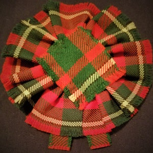 May include: A red, green, and white plaid fabric bow with fringed edges. The bow is made of a woven fabric with a plaid pattern.