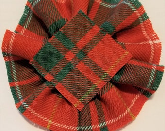 May include: A red, green, and white plaid fabric bow with a square center piece. The bow is made of a woven fabric with a fringed edge.