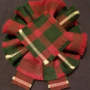 Puede incluir: Un lazo de tela a cuadros rojo, verde y blanco. El lazo está hecho de un material suave y peludo y tiene un borde con flecos.