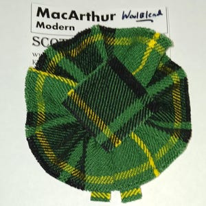 Op de afbeelding: Een groene, zwarte en gele geruite stof met de tekst "MacArthur Woolblend Modern" op een witte achtergrond.