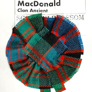 Könnte beinhalten: Ein rundes, plissiertes Stoffelement in einem Tartanmuster aus Blau, Grün, Rot und Schwarz. Der Artikel hat ein Etikett mit der Aufschrift "MacDonald Clan Ancient". Der Stoff ist zu einer kreisförmigen Form mit gewelltem Rand gefaltet.