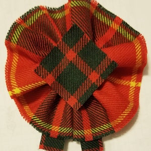 May include: A red, green, and yellow plaid fabric bow with a square center. The bow is made of a soft, woven fabric and has a fringed edge.