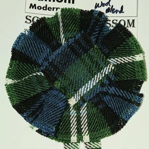 Könnte beinhalten: Ein schottischer Kilt-Pin aus grünem, blauem und weißem kariertem Stoff mit dem Text "Lamont Modern Scottish Blossom Wood Blend" auf einem weißen Etikett.