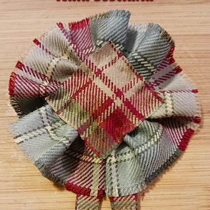 May include: A handmade plaid fabric flower with a central square and layered petals. The flower features a mix of red, green, and gray tones, with the text "Auld Scotland" in red at the top. The fabric has a fringed edge.
