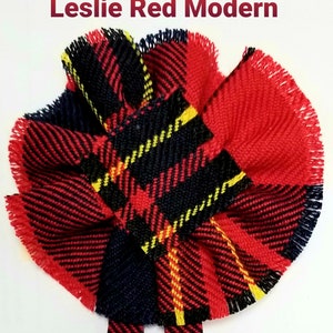 May include: A red, black, and yellow plaid fabric flower with frayed edges. The fabric is folded and arranged to create a flower shape. The flower is about 4 inches in diameter.